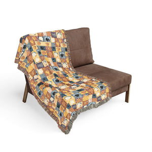 A brown upholstered lounge chair on a white background with a colorful patterned throw blanket draped over it. The blanket features an abstract swirling design in yellow, blue, orange, and purple tones with fringed edges.