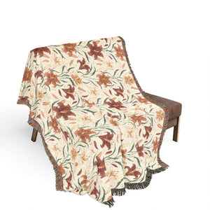 A fringed throw blanket draped over a brown armchair, featuring a botanical pattern of brown and orange flowers with green leaves on a cream background.