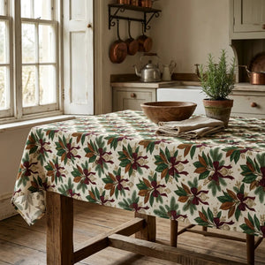 A square tablecloth with a repeating floral pattern in green, burgundy, and brown on a cream background, shown draped over a table with light wood legs against a white background.