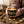A person's hands holding a small black metal candle tin with a gold and green lotus pattern, lit with a single wick, against a plain white background.