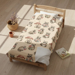 A wooden single bed in a sunlit room, covered with a cream-colored blanket printed all over with raccoons and mushroom motifs. A woven basket with dried flowers, a small orange mug, and a children's book sit nearby on the light wood floor.